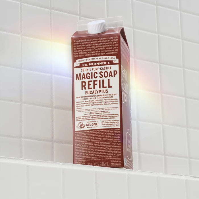 A brown carton labeled MAGIC SOAP REFILL sits on a white tiled shelf, illuminated by a rainbow reflection. The packaging promotes Dr. Bronners natural, eco friendly, organic soap benefits [32 oz]