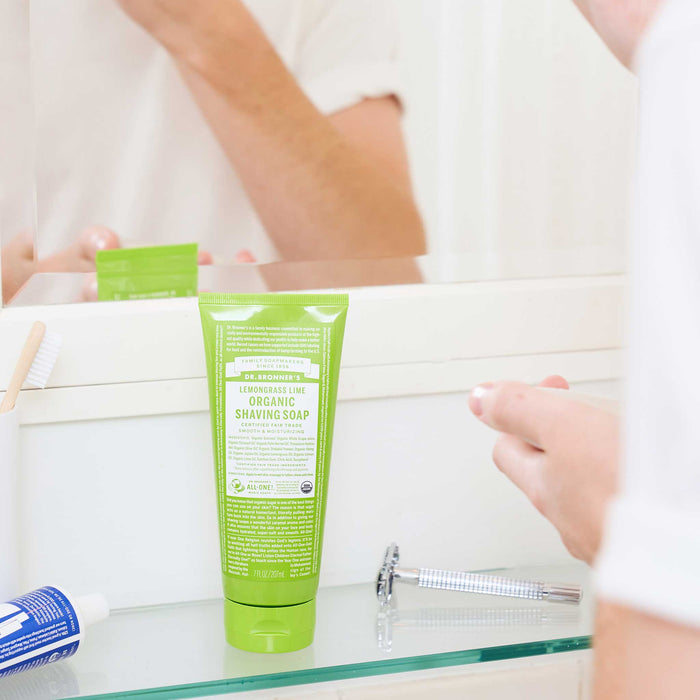 A person handles shaving cream next to a mirror. In front is a green tube labeled Dr. Bronners Lemongrass Lime Organic Shaving Soap amid other grooming items. The setting is a bathroom [7 oz]