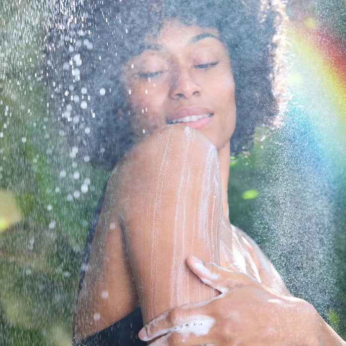 A person enjoys an outdoor shower, applying soap to their arm, surrounded by a lush, green garden with a subtle rainbow in the background, evoking a refreshing, natural experience [5 oz]