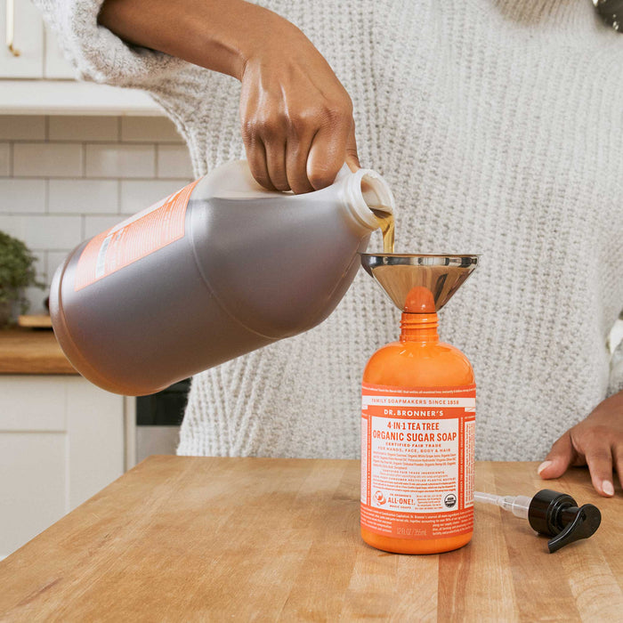 A person pours Dr. Bronners Tea Tree Organic Sugar Soap from a large container into a smaller bottle using a funnel in a bright kitchen. DR. BRONNERS ORGANIC SUGAR SOAP  [12 oz]