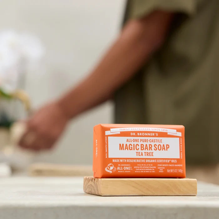 A bar of Dr. Bronners Magic Bar Soap in tea tree variant sits on a wooden dish. In the background, a person reaches towards a blurred table, suggesting a home setting. DR. BRONNERS ALL ONE PURE CASTILE MAGIC BAR SOAP TEA TREE MADE WITH REGENERATIVE ORGANIC CERTIFIED OILS [5 oz]
