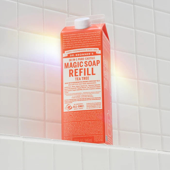 Dr. Bronners 18 in 1 Pure Castile Magic Soap Refill in Tea Tree scent sits upright on a white tiled shelf. The packaging highlights eco friendly and fair trade qualities, emphasizing sustainable practices [32 oz]